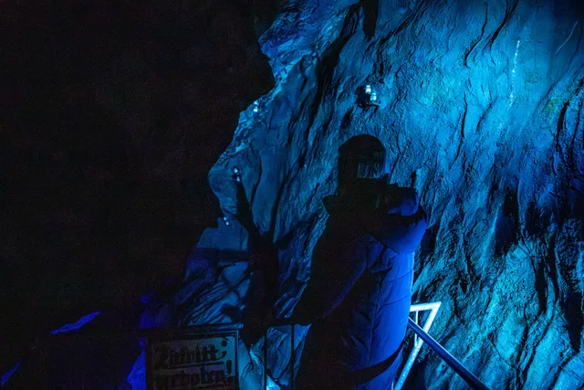 Person mit Rucksack fotografiert in einer bläulich beleuchteten Höhle, Felswände und Geländer.