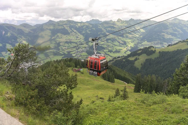 Rote Seilbahn-Gondel schwebt über grüne Alpenlandschaft mit Bergen im Hintergrund.