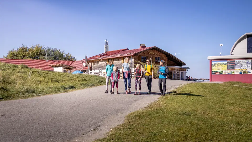 Gruppe von sieben Wanderern spaziert auf einem Weg neben Gebäuden bei sonnigem Wetter.