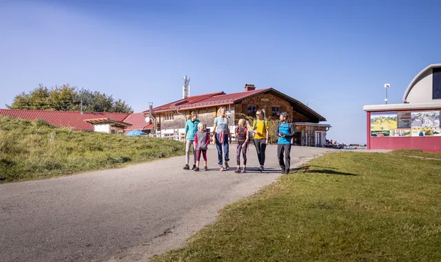 Gruppe von sieben Wanderern spaziert auf einem Weg neben Gebäuden bei sonnigem Wetter.