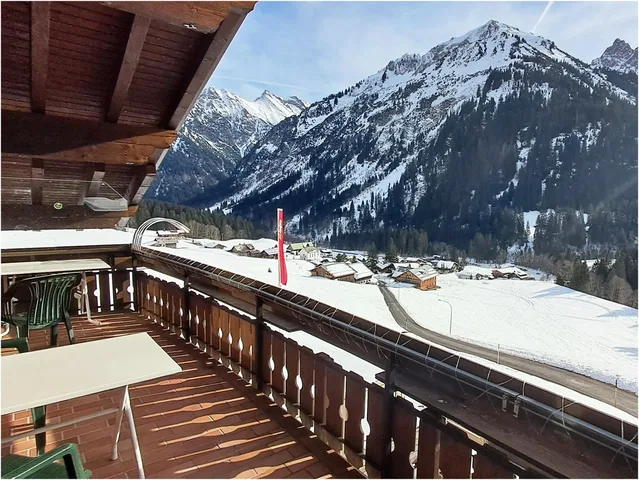 Balkon mit Tisch und Stühlen, verschneite Berglandschaft und Chalets im Tal.