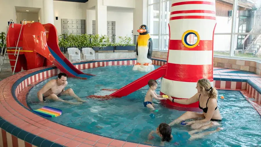 Familie im Hallenbad-Kinderbecken mit roter Rutsche und Pinguin-Dekoration.