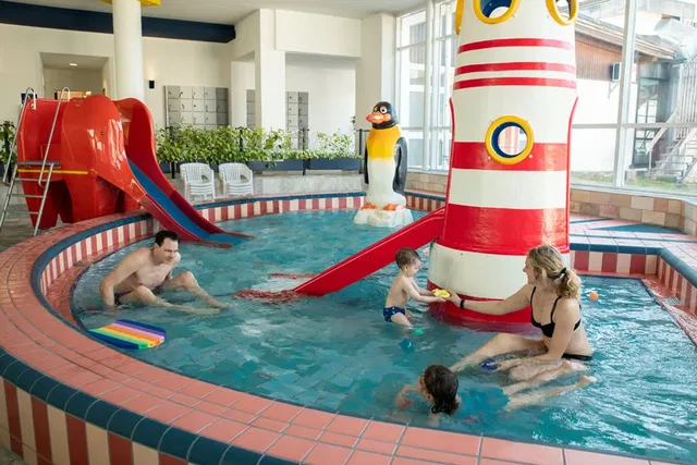 Familie im Hallenbad-Kinderbecken mit roter Rutsche und Pinguin-Dekoration.