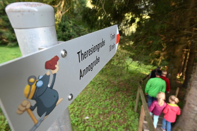 Familie wandert auf dem Weg zu Theresien- und Annagrube, im Vordergrund ein Wegweiser mit Knappenfigur.