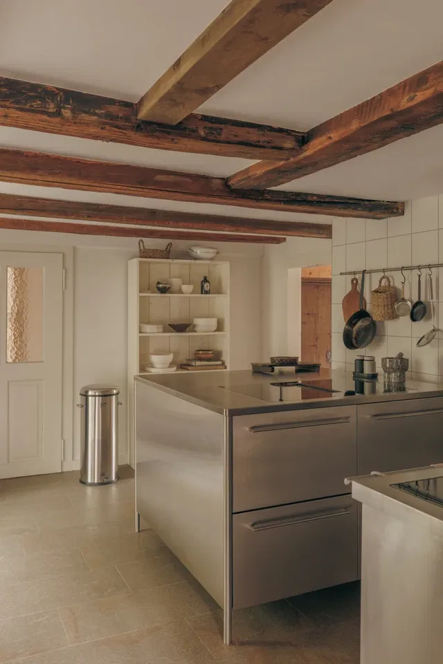 Küche mit sichtbaren Holzbalken an der Decke, Edelstahlinsel und offener Regalwand.