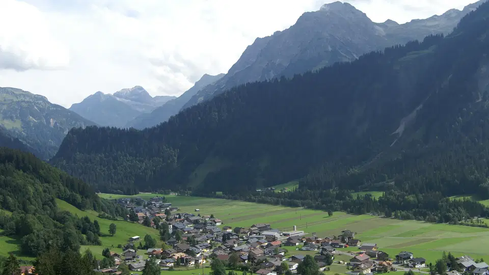 Landschaft mit Talort, grünem Feldland und Dorf am Waldrand vor Bergen.