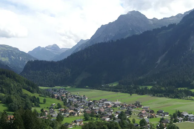 Landschaft mit Talort, grünem Feldland und Dorf am Waldrand vor Bergen.