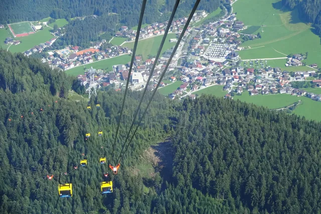 Seilbahngondeln über einem Wald mit Siedlung und Wiesen im Hintergrund