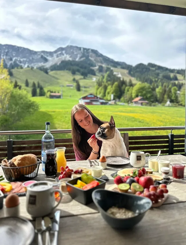 Frau mit Hund am Frühstückstisch im Freien vor Berglandschaft