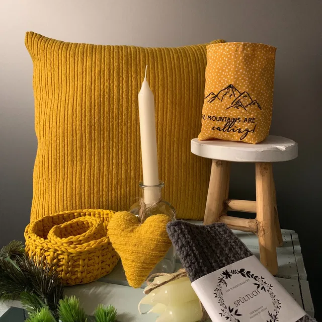 Gelbes Kissen, Kerze und gestricktes Herz neben einem Körbchen mit Spültuch.