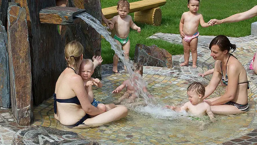 Gruppe Erwachsener und Kleinkinder im Outdoor-Kinderbecken, Wasser aus einer Steinquelle sprudelt.
