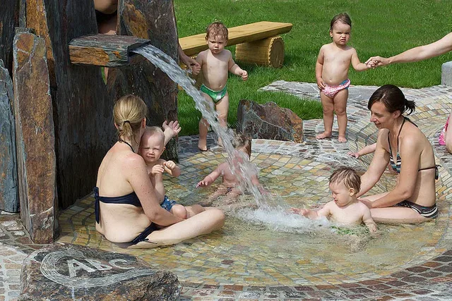Gruppe Erwachsener und Kleinkinder im Outdoor-Kinderbecken, Wasser aus einer Steinquelle sprudelt.