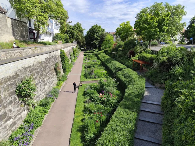 Länglicher Gartenpfad flankiert von Steinmauern und hohen Hecken, mit Personen am Weg.