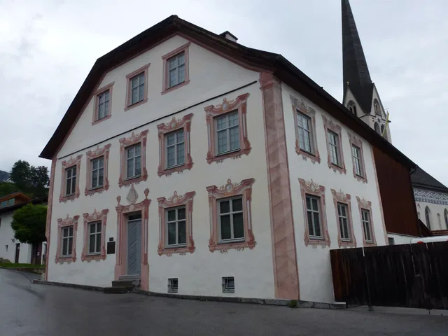 Historisches weißes Haus mit rosa Fassadendekor und vielen Fenstern; Kirchturm im Hintergrund.