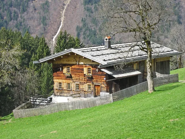 Rustikale Holzhütte auf grünem Hügel, von Wald und Zaun umgeben.