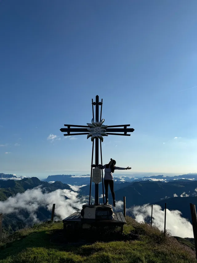 Person neben großem Kreuz auf Berggipfel mit Blick auf Wolken und Bergkette.