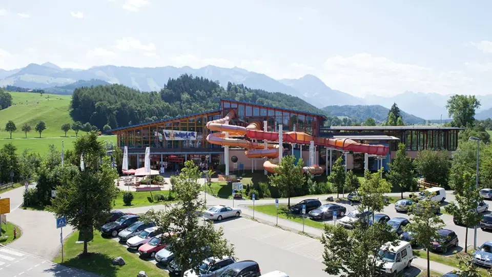 Wasserpark mit rotem/orangem Rutschen-Complex, Gebäude und Parkplatz mit Bäumen, Alpen im Fernblick.