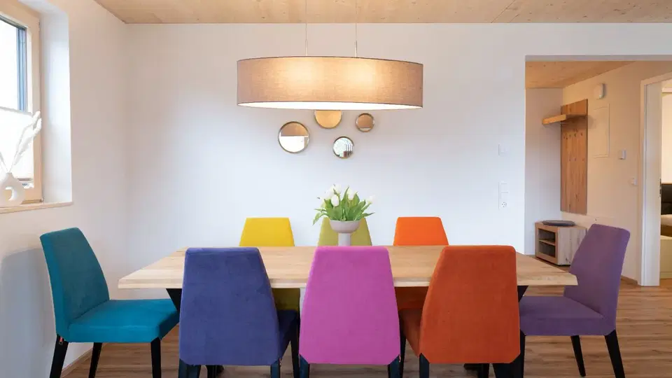 Helles Esszimmer mit Holz-Tisch und bunten Stühlen um ihn herum, großer Lampenschirm darüber