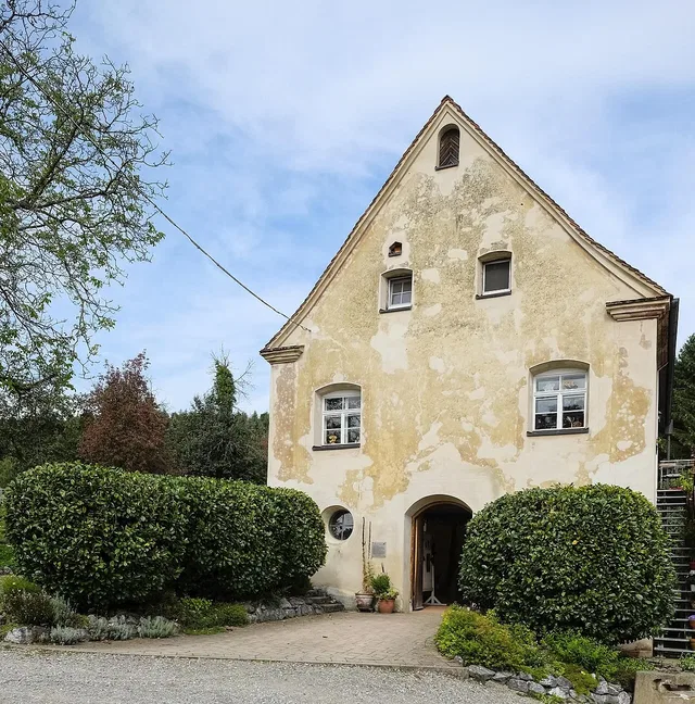 Historisches gelb verputztes Haus mit spitzem Dach und offener Eingangstür, von Sträuchern umrahmt.