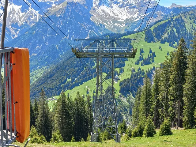 Liftturm im Bergland mit Seilbahnkabeln, grüne Wälder und schneebedeckte Berge im Hintergrund.