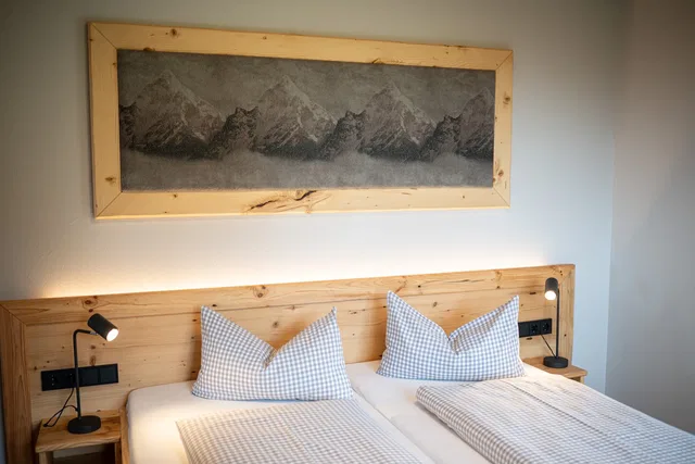 Zweibettzimmer mit Holzbettkopfteil, zwei Checkkissen, Bergbild über dem Bett.