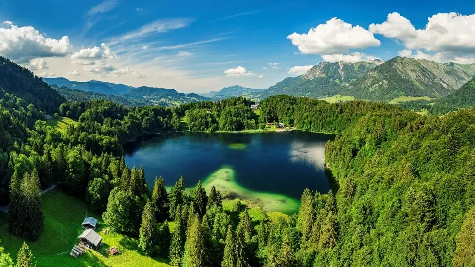 Landschaft mit einem stillen Bergsee, umgeben von Wald und Bergen unter blauem Himmel.