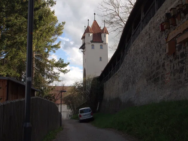 Gasse führt zu weißem Turm mit roten Türmen, von Mauern und Bäumen umgeben.