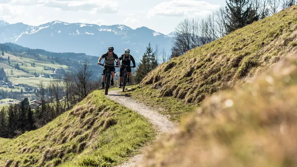 Zwei Mountainbiker folgen einem schmalen Pfad am Hang mit Berglandschaft im Hintergrund.