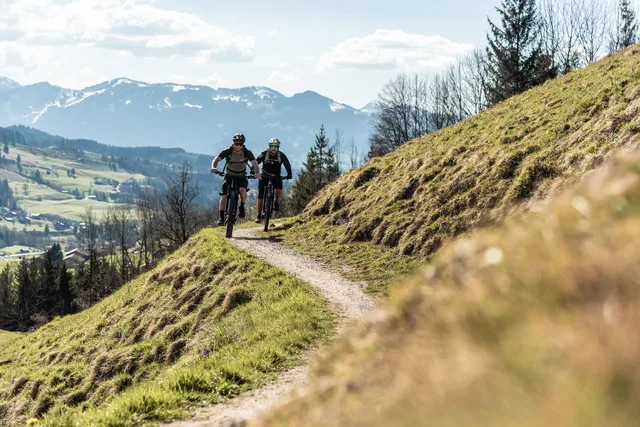 Zwei Mountainbiker folgen einem schmalen Pfad am Hang mit Berglandschaft im Hintergrund.