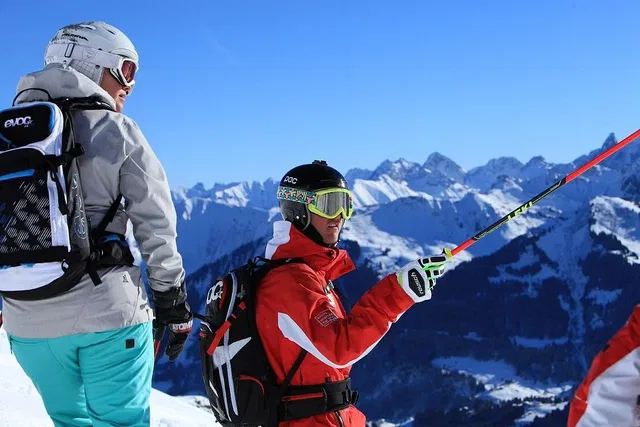 Zwei Wintersportler in Berglandschaft; einer in rotem Mantel, einer grauen Jacke mit Helm