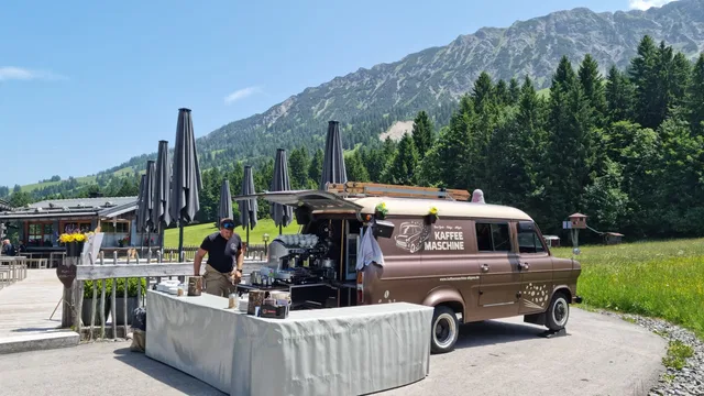 Brauner Kaffee‑Van mit offener Theke im Freien, Barista bereitet Getränke, Berglandschaft im Hinterg