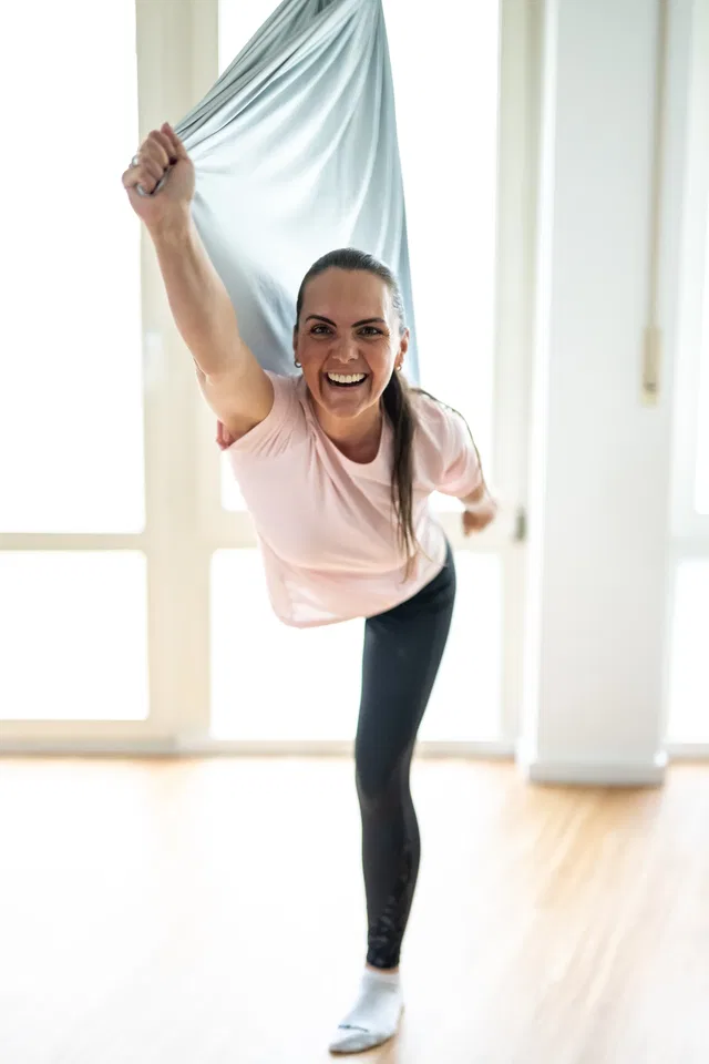 Frau trainiert mit Aerial-Silk im hellen Studio, hebt den Arm und lächelt.