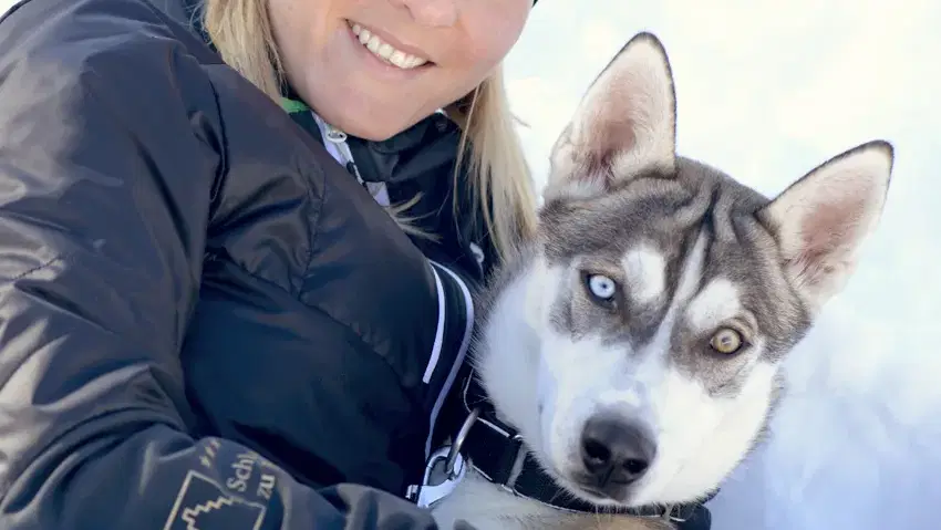 Frau in Winterkleidung mit Skibrille hält einen Husky im Schnee.