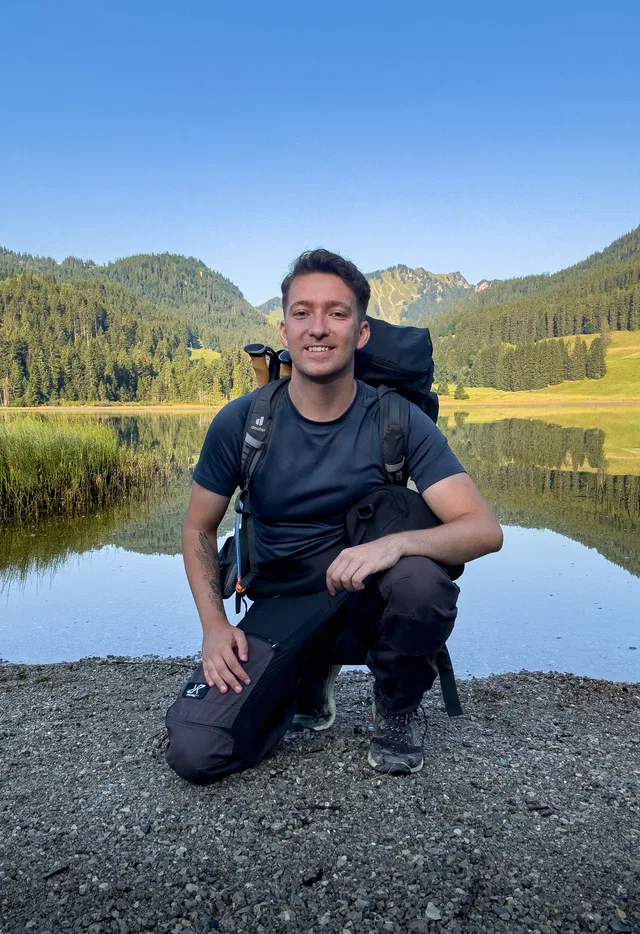 Junger Mann mit Rucksack kniet am Ufer eines klaren Sees in einer bergigen Waldlandschaft.