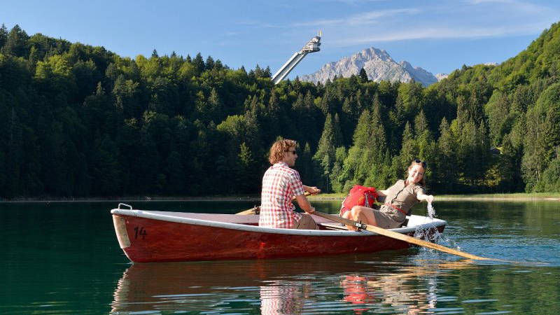 Zwei Personen in einem Ruderboot auf dem Freibergsee vor Wald und Skiflugschanze.