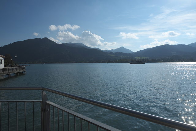 Blick vom Ufer in Tegernsee über den See auf ein Ausflugsschiff und die Berge.