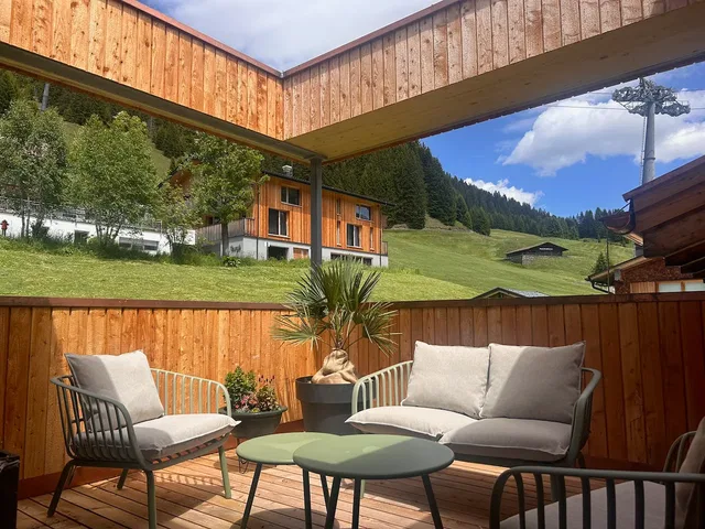 Holzterrasse mit Lounge-Möbeln, Topfpflanzen und Blick auf Hügel und Gebäude.