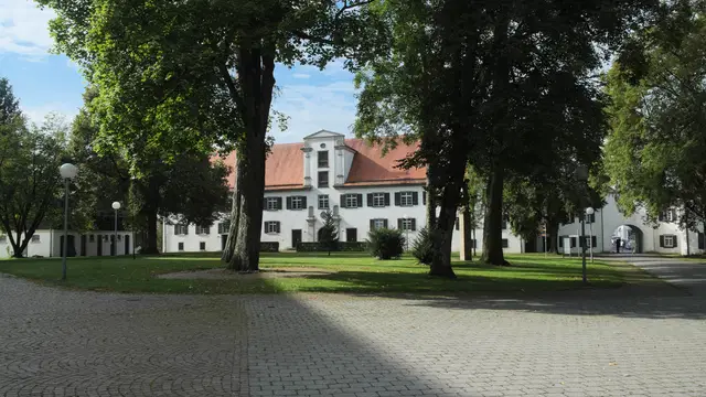 Fassade des Klosters Schussenried hinter großen Bäumen mit gepflastertem Vorplatz. Ortsmotiv zur Ausflugsplanung.
