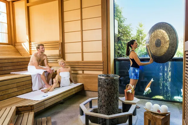 Zwei Personen in Saunatücher sitzen auf Holzbänken, eine Frau schlägt Gong in einem Spa