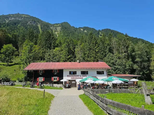 Restaurant mit rotem Dach vor Waldhügeln, Außenbereich mit Tischen und grünen Sonnenschirmen