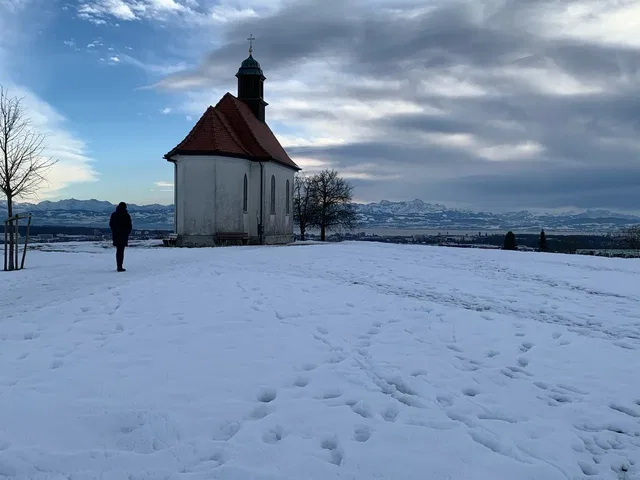 Kleine Kapelle mit rotem Dach auf verschneiter Wiese; eine Person geht im Vordergrund.