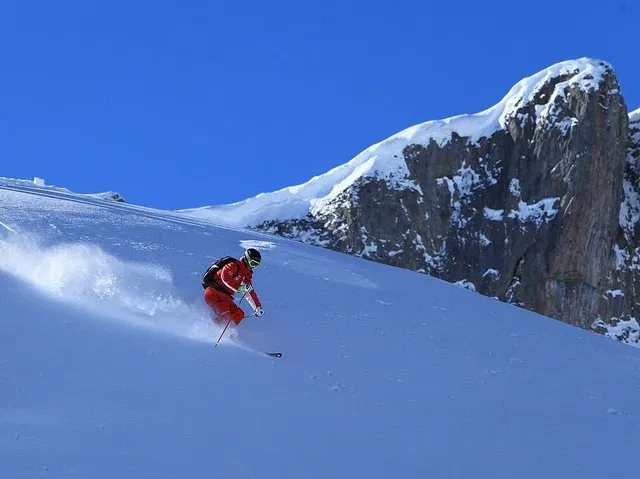 Skifahrer in roter Kleidung fährt eine verschneite Abfahrt vor einem felsigen Berguntergrund