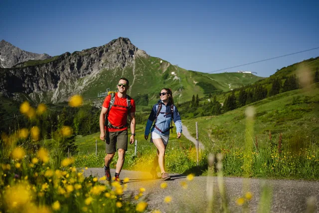 Paar wandert auf einer Alpenwiese mit Bergen im Hintergrund, Gelbblumen im Vordergrund.