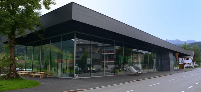 Modernes Glasgebäude mit schwarzem Flachdach und großen Fenstern, von Bäumen umgeben.