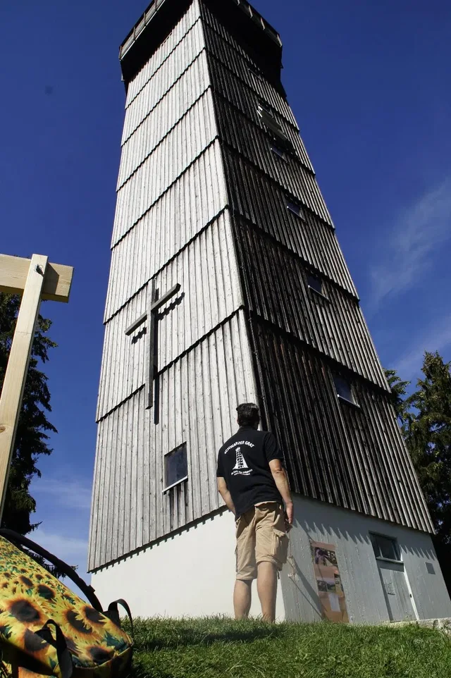 Hoher Holzturm aus vertikalen Bretterlagen, Mann steht am Fuß davor und schaut ihn an.