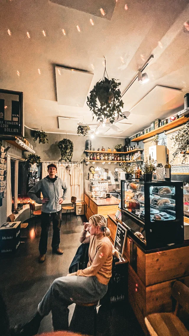 Zwei Personen in einem gemütlichen Café: Ein Mann steht links, eine Frau sitzt rechts am Tisch.