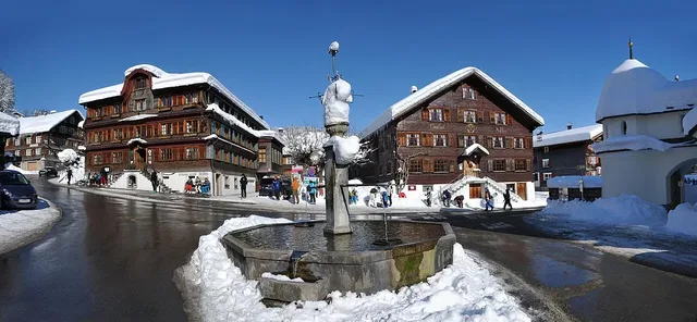Winteransicht eines Dorfplatzes mit Brunnen, Holzhäusern und Schnee.