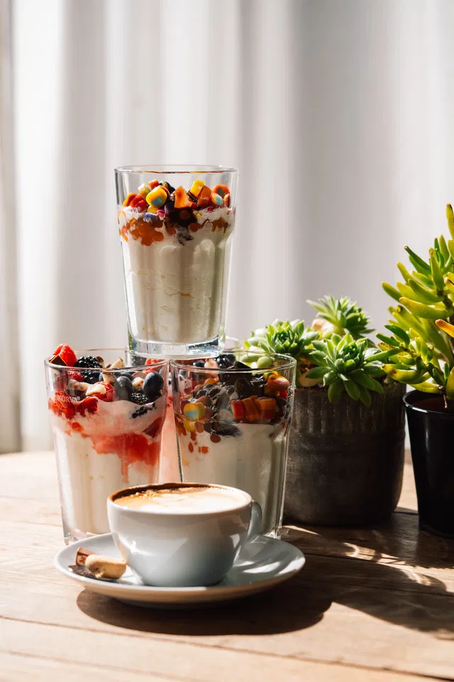 Mehrere Gläser mit Joghurt-Dessert und Obst, daneben eine Kaffeetasse; Sukkulenten im Hintergrund.