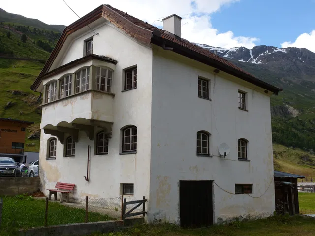 Weißes zweistöckiges Haus mit Balkon vor Berglandschaft und Tal.