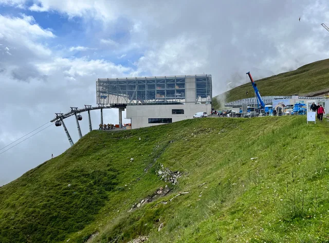 Moderne Bergstation an einer Gebirgskante mit Seilbahnanlage und Besucherbereich auf grünem Gras
